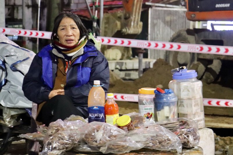 Cụ bà bán hàng rong trên phố, dù không mưa nhưng bà cũng phải khoác thêm một chiếc áo mưa để giữ ấm, vì không đủ áo quần để mặc trong thời tiết giá lạnh này.